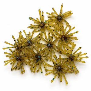 May include: A collection of eight translucent olive-green fishing lures, each featuring a central hub with numerous radiating flexible appendages. The lures are speckled with small black and metallic blue glitter throughout.