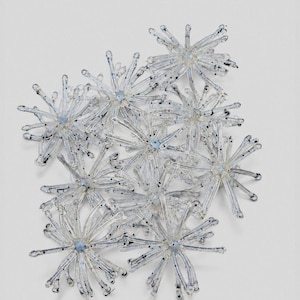 May include: A cluster of clear glass starburst ornaments, each featuring a central light blue dot and radiating thin, translucent spokes with small dark speckles at the tips, arranged in a dense, overlapping formation on a neutral background.