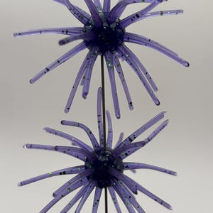 May include: Two purple, spiky decorative ornaments. Each ornament has a dark purple center with radiating, slender, translucent purple arms. The arms are dotted with green glitter. The ornaments are attached to a thin, dark metal rod.