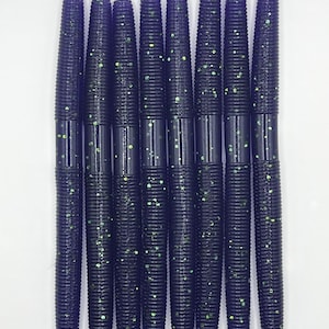 May include: A collection of nine dark purple fishing lures with a ribbed design. The lures are speckled with green glitter, and are arranged vertically against a white background. These soft plastic lures are designed for attracting fish.