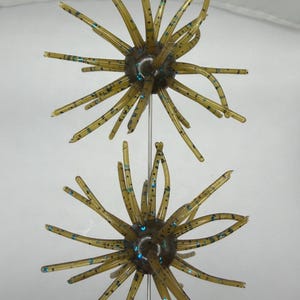 May include: Two olive-green fishing lures with multiple radiating appendages and blue speckles. The lures have a central, dark-colored body. The image is taken against a white background, showcasing the detailed design of the artificial bait.