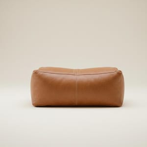 May include: A rectangular, brown leather ottoman with rounded edges. The ottoman has a stitched seam detail down the center and is set against a neutral background. This piece of furniture is designed for seating or footrest.