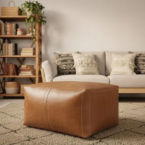 May include: A square, brown leather ottoman with visible stitching details. The ottoman is placed on a patterned rug in front of a white sofa with decorative pillows. A wooden bookshelf with books and plants is in the background.