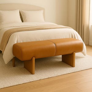 Puede incluir: Banco rectangular moderno con asiento de cuero marrón claro y patas de madera. El banco está colocado al pie de una cama con edredón y almohadas color crema. La habitación tiene una paleta de colores neutros.
