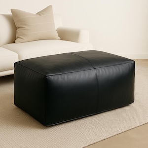 May include: A large, square black ottoman with stitched detailing. The ottoman is made of a smooth, leather-like material and sits on a neutral-colored rug. A cream-colored sofa and a striped pillow are in the background.