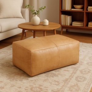 May include: A rectangular, tan leather ottoman with stitched detailing. The ottoman is placed on a patterned rug in a living room setting. A round wooden coffee table and a white sofa are visible in the background.