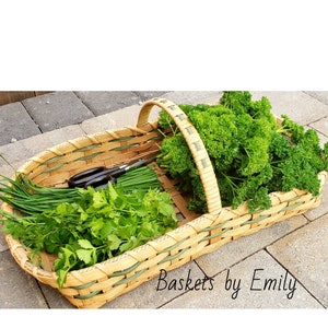 May include: A woven basket with a handle, filled with fresh green herbs, including parsley and chives. The basket is made of natural materials and has a light brown color. The text "Baskets by Emily" is visible in the bottom right corner of the image.