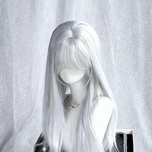 Puede incluir: Una peluca larga de color gris plateado con flequillo recto, peinada sobre una cabeza de maniquí. La peluca presenta capas largas y rectas y una línea de cabello de aspecto natural. Se ve un collar plateado. El fondo es una tela blanca texturizada.