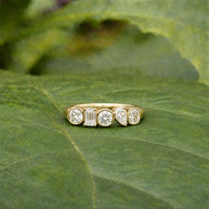 Peut inclure: Une bague en or ornée de cinq pierres précieuses transparentes de formes variées : rondes, taille émeraude, rondes, poire et rondes. La bague est posée sur une feuille verte, créant un contraste avec le fond naturel.