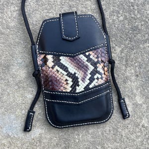 Op de afbeelding: Zwarte leren crossbody telefoontas met een slangenprint. De tas heeft witte stiksels en een zwart koord. De slangenprint heeft bruine, beige en paarse tinten.