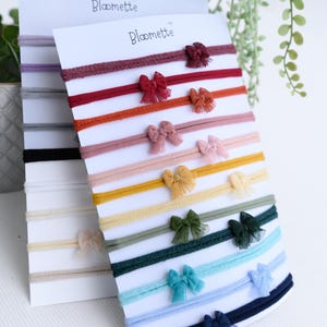 Puede incluir: Un conjunto de diademas para bebés en varios colores, cada una adornada con un pequeño lazo. Las diademas se exhiben en tarjetas blancas con el nombre de la marca "Bloomette" impreso en la parte superior. Colores: burdeos, rosa, amarillo, verde y azul.