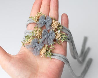 May include: A set of three thin grey elastic headbands, each featuring a small grey tulle bow and clusters of dried beige flowers with green stems. The headbands are arranged in a stack, showcasing the delicate floral and fabric details.