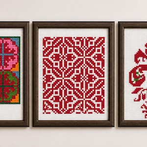May include: Three framed cross-stitch artworks. The left frame features a colorful geometric design with pink, green, and orange elements. The center frame displays a red and white geometric pattern. The right frame showcases a red floral design.