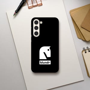 May include: Black phone case with a white horse head logo and the word "blundr" in a white rectangle. The phone case has a camera module with three lenses. A pen and paper are in the background.
