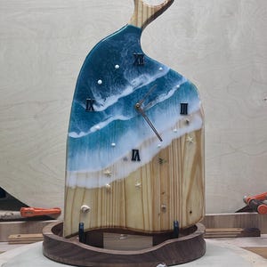 May include: A wooden clock with a beach-themed design. The top portion features blue and white resin waves, while the bottom has a light wood grain appearance. Roman numerals mark the hours. The clock is supported by a wooden base.