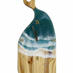 May include: Wooden wall clock with a beach-themed design. The clock face features a resin ocean scene with blue and white waves, a sandy beach, and gold Roman numerals. The handle is made of wood.