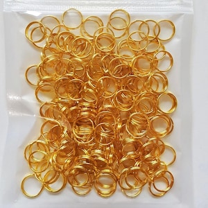 May include: A transparent bag containing a large quantity of small, shiny, gold-coloured metal rings. The rings are closely packed, forming a dense cluster. The rings appear to be made of a smooth, reflective material, and are all the same size and shape.