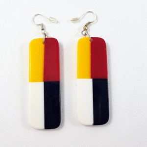 May include: A pair of long rectangular earrings with a geometric design. The earrings are made of yellow, red, white, and black resin. The earrings have silver hooks.