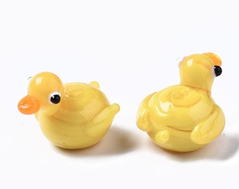 May include: Two small yellow glass duck figurines with black eyes. The ducks have a swirl pattern and are sitting on a white surface.