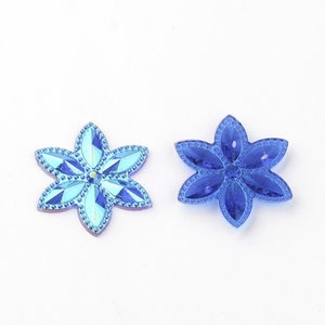 May include: Two blue flower-shaped rhinestones with a faceted design. The rhinestone on the left has a lighter blue color and a more iridescent finish. The rhinestone on the right is a darker blue color.