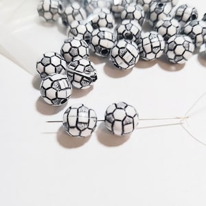 May include: White and black soccer ball-shaped beads. The beads are strung on a needle with white thread. The beads are made of a glossy material and have a detailed soccer ball design. A clear plastic bag is in the background.