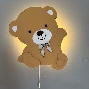 Può includere: Una lampada da parete a forma di orsetto marrone con fiocco e dettagli bianchi. La luce è accesa, illuminando lo spazio dietro l'orso. Un cordoncino bianco pende sotto.