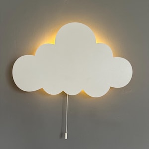 May include: White cloud-shaped wall light fixture with a warm yellow glow emanating from behind the cloud. The light has a pull-chain switch and is mounted on a gray wall. A decorative and functional lighting solution.