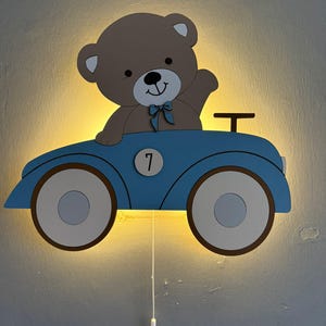 Puede incluir: Una lámpara de pared LED con forma de oso de peluche marrón conduciendo un coche azul. El oso lleva una pajarita azul y el coche tiene un círculo blanco con el número 7 en el lateral. Un cordón blanco cuelga de la parte inferior del coche.
