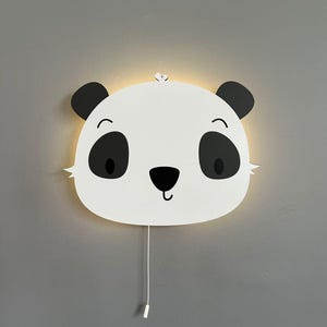 Könnte beinhalten: Eine weiße Wandleuchte in Pandaform mit schwarzen Akzenten für Augen, Ohren und Nase. Das Licht hat ein lächelndes Gesicht und eine Zugschnur. An einer grauen Wand montiert.