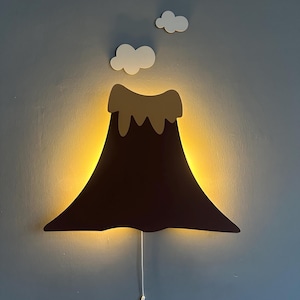 Puede incluir: Una lámpara de pared con forma de volcán, con un cuerpo marrón oscuro y una parte superior marrón claro, que emite un brillo amarillo cálido. Dos recortes de nubes blancas están encima del volcán. La lámpara está montada en una pared azul grisáceo y tiene un cordón blanco.