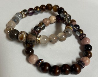 Könnte beinhalten: Zwei Armbänder mit Perlen aus Naturstein in Braun-, Rosa- und transparenten Farbtönen. Die Armbänder haben goldene und kristallene Akzente. Die Perlen variieren in Größe und Textur und schaffen ein einzigartiges Accessoire.
