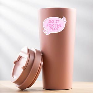 May include: A pink travel mug with a matching lid is on a wooden surface. A pink sticker on the mug says "DO IT FOR THE PLOT" in white text. The mug is designed for hot or cold drinks. The mug's height is approximately 20 cm.