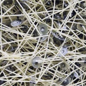 May include: Overhead shot of a pile of candle wicks and wick holders. The wicks are a light beige color, and the metal wick holders are silver. The wicks are arranged in a tangled, crisscross pattern.