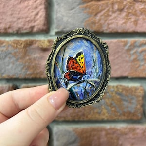 Puede incluir: Un broche de forma ovalada que presenta una pintura al óleo en miniatura de una mariposa naranja y negra posada sobre briznas de hierba. La obra está encerrada en un marco metálico oscuro y ornamentado con un acabado texturizado antiguo.