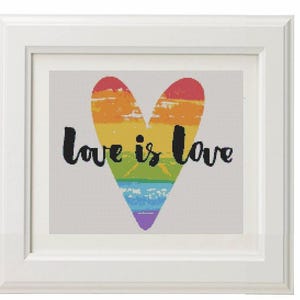 May include: A framed artwork features a rainbow-colored heart with the words "love is love" in black script. The heart design includes bands of red, orange, yellow, green, blue, and purple. The artwork is set against a white background and framed in a white wooden frame.
