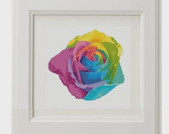 May include: A framed cross-stitch art piece featuring a rainbow rose. The rose's petals transition through shades of pink, blue, green, and yellow. The artwork is set against a white background within a white frame.