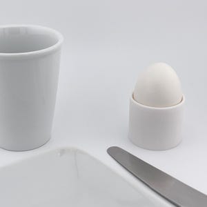 Könnte beinhalten: Ein weißer Keramikbecher, Eierbecher, Teller und ein Edelstahlmesser sind auf einer weißen Oberfläche angeordnet. Der Eierbecher enthält ein weißes Ei. Die Artikel sind vor einem weißen Hintergrund platziert.