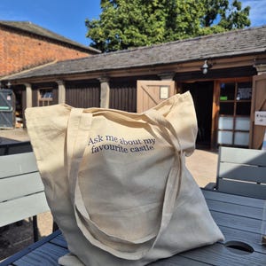 May include: A cream-coloured canvas tote bag with the text "Ask me about my favourite castle." The bag has long straps and is on a grey table. A building and a tree are visible in the background.
