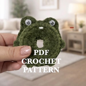 May include: A dark green crocheted frog with large googly eyes and a white accent. The frog is donut-shaped with the text "PDF CROCHET PATTERN" in white. The frog is held in a hand.