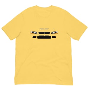 Peut inclure: T-shirt jaune avec un graphisme représentant l'avant d'une voiture. Le design comprend des phares, une calandre et d'autres détails en noir, orange et blanc. Le t-shirt est fait d'une matière douce.