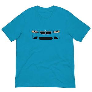 May include: A turquoise t-shirt with a graphic of a car's front. The design features headlights, a grille, and a bumper in black and yellow against a blue backdrop. The shirt is made of a soft material.