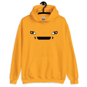 Könnte beinhalten: Senfgelber Kapuzenpullover mit einem Cartoon-Gesicht-Design. Das Design zeigt schwarz-weiße Augen, einen schwarzen Mund und eine kleine horizontale Linie über dem Mund. Der Hoodie hat eine Vordertasche und eine Kapuze mit Kordelzug.