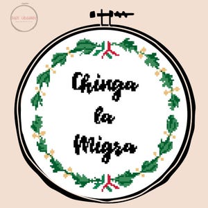 May include: A cross-stitch embroidery hoop with the text "Chinga la Migra" in black, surrounded by a green and gold leaf design. The hoop is set against a light peach background, with a small logo in the upper left corner.