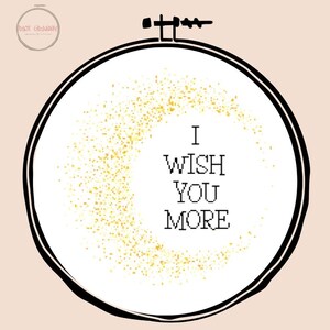 Op de afbeelding: Een kruissteekontwerp in een ronde borduurring. Het ontwerp toont de tekst "I WISH YOU MORE" in zwart, met een gouden glitterachtige achtergrond. De ring is zwart met een lichtroze achtergrond.
