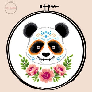 May include: A cross-stitch embroidery hoop featuring a panda bear design. The panda has black ears and eye markings, orange accents, and a sugar skull-inspired face. Pink flowers and green leaves adorn the bottom. The hoop is black and the background is white.