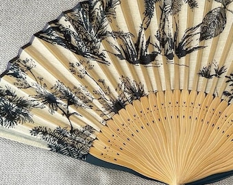 Op de afbeelding: Een vouwbare handwaaier met een beige stoffen blad bedrukt met een zwarte inktlandschapsillustratie van bomen en gebladerte. De waaier heeft lichtgekleurde houten ribben en een donker handvat, afgewerkt met een kleine decoratieve kwast.