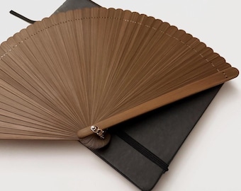 Op de afbeelding: Een bruine vouwbare handwaaier met een geschulpte rand en een zilverkleurige metalen klinknagel op het draaipunt, rustend op een zwart rechthoekig notitieboek met een elastische sluitband en een zwart lint als bladwijzer aan de bovenkant.