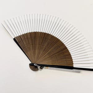 Könnte beinhalten: Ein handgeführter Faltfächer mit einem einfarbig weißen Papierblatt, das an einem dunkelbraunen Holzrahmen befestigt ist. Der Griff ist schwarz und verfügt über einen kleinen silbernen Metallring mit einer langen, dünnen, hellgrauen Quaste.