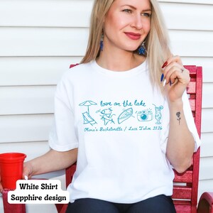 以下が含まれることがあります： 白いTシャツ。サファイアブルーのデザインで、「love on the lake」の文字と、パラソル、ラウンジチェア、人々、ボート、カメラ、カクテルのイラストが描かれています。「Olivia's Bachelorette / Lake Tahoe 2026」の文字も含まれています。