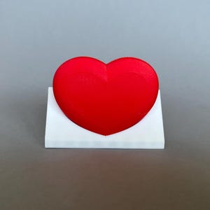 Può includere: Un oggetto a forma di cuore rosso vivo poggia su una base rettangolare bianca. Il cuore è liscio e pieno, con una leggera rientranza nella parte superiore. La base ha un design pulito e semplice, che offre contrasto. L'immagine è un primo piano.